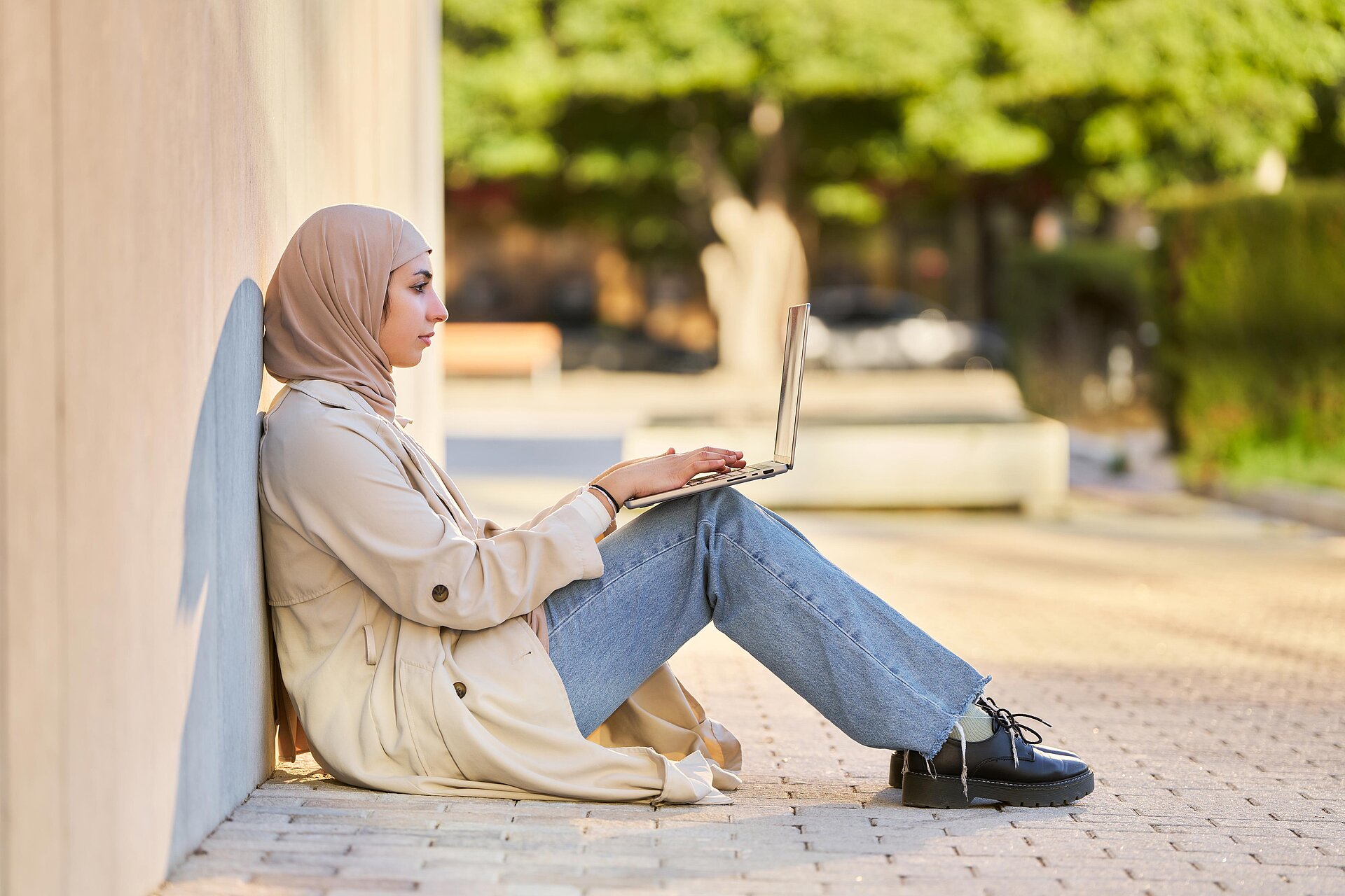 Girl wearing a hijab sitting outside on a wall with a laptop on her knees.